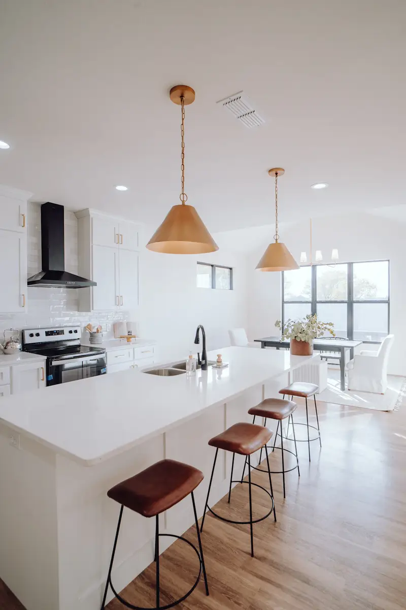 Cocina nueva en blanco con isla, lámparas colgantes y taburetes.